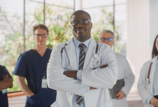 Team Portrait of a Female and Male Successful Diverse Medical Healthcare Professionals Standing as a Group in a Modern Hospital Office. African American Doctor Standing Closer to Camera