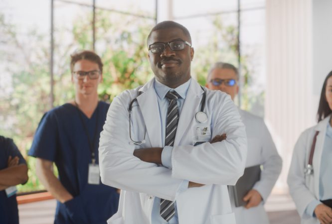 Team Portrait of a Female and Male Successful Diverse Medical Healthcare Professionals Standing as a Group in a Modern Hospital Office. African American Doctor Standing Closer to Camera